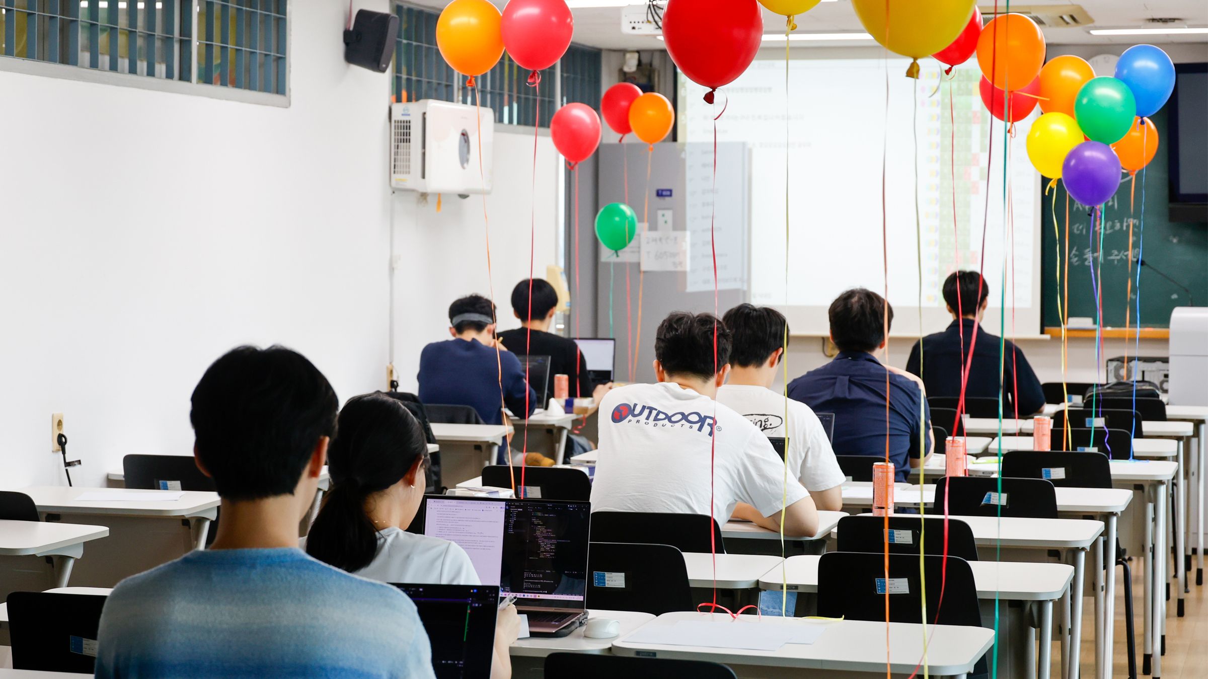 교실 앞쪽 화면을 향해 앉은 참가자들의 뒷모습과, 천장까지 떠오른 풍선 다발.